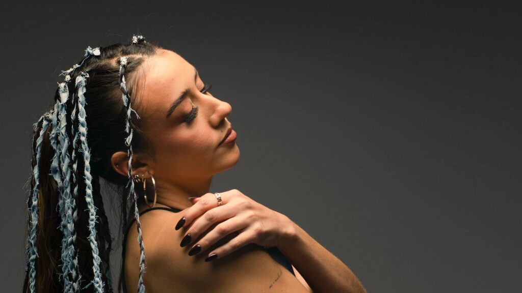 Lua Lacruz, artista chilena emergente, en photoshoot promocional para su nuevo single "Autosabotaje". Lua está en un fondo gris oscuro, ladeada hacia la izquierda y luce trenzas en su cabello